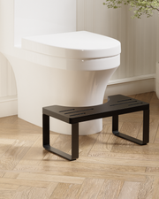 A black metal toilet step stool placed in front of a white toilet on a light wood herringbone floor in a bathroom setting.