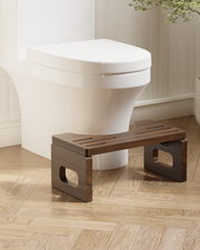 A dark brown wooden toilet step stool placed in front of a white toilet on a light wood herringbone floor.