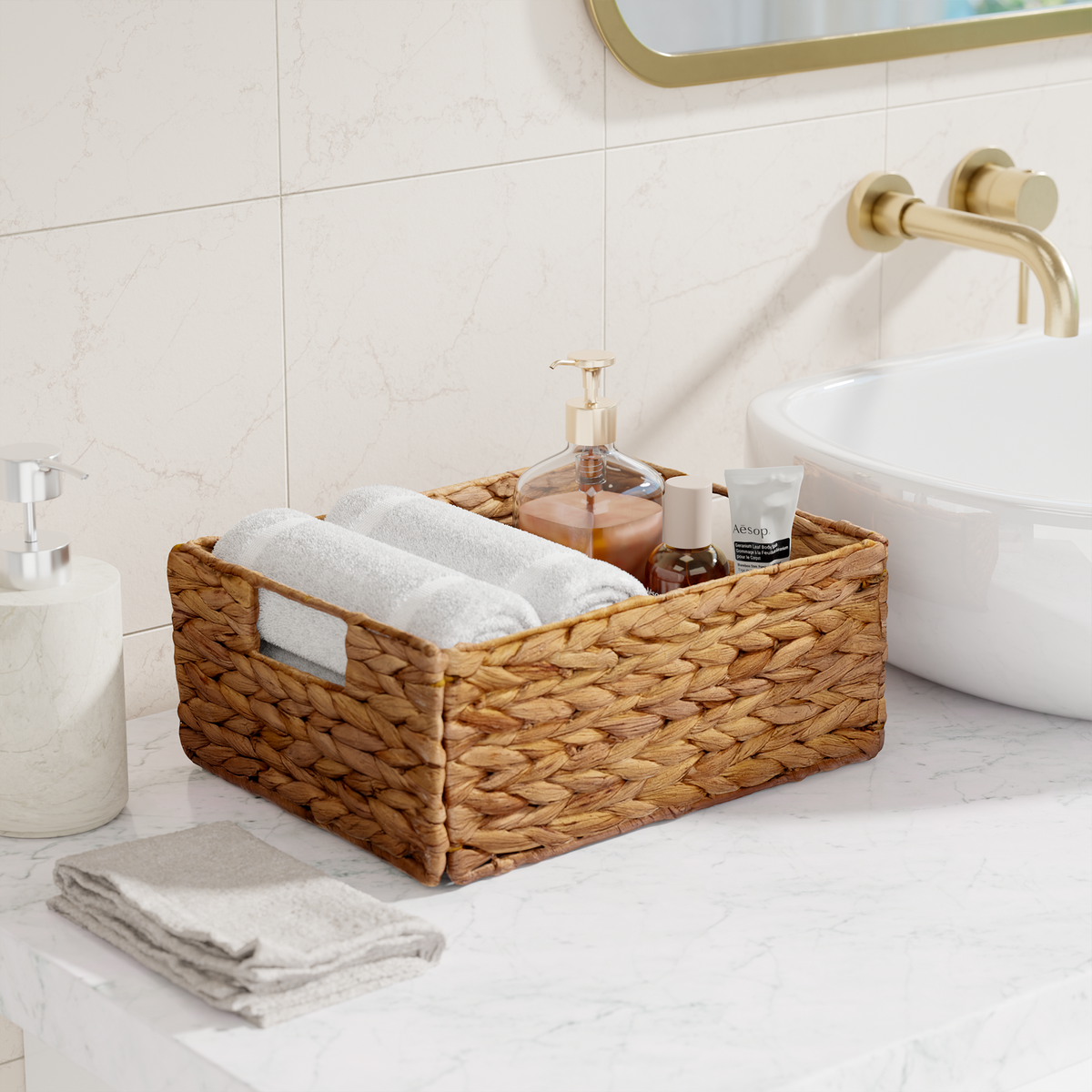 A woven rectangular storage basket with cutout handles sits on a bathroom counter, holding rolled white towels and small toiletries. The basket is styled in a clean, modern bathroom setting with a marble sink, gold faucet, and cream tiles.