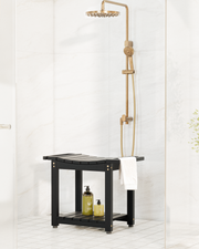 A modern shower setup featuring a black wooden bench with a lower shelf, a brass shower head and handheld hose, and a white towel, set against a white marble-tiled bathroom wall.