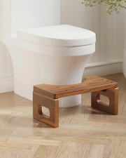 A wooden bathroom step stool with cutout legs, positioned in front of a white toilet on a light wood herringbone floor.
