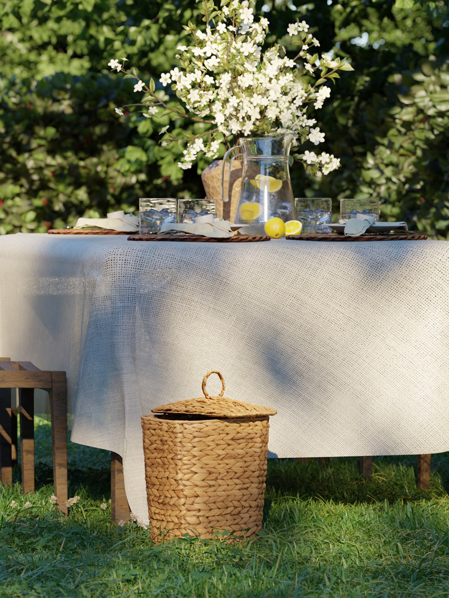 Outdoor setting with a table covered by a white tablecloth, a vase with flowers, and a woven basket on a grassy background.